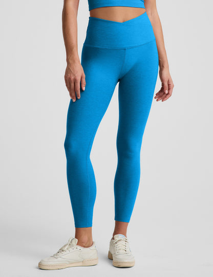 Woman wearing a blue cropped tank top and blue high waisted midi leggings with a v front waistband against a plain background.