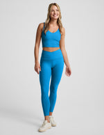 Woman wearing a blue cropped tank top and blue high waisted midi leggings with a v front waistband against a plain background.