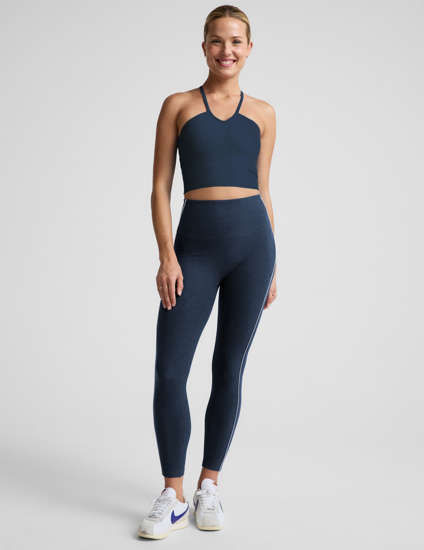 Woman wearing a navy blue cropped tank top and midi leggings with white contrast piping on the sides against a plain background.
