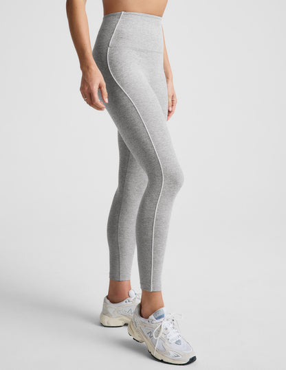 Woman wearing a gray sports bra and leggings with white contrast piping on a plain background.