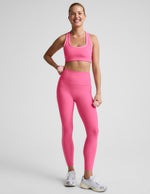 Woman wearing a pink bra and pink midi leggings with white contrast piping on the sides against a plain background.