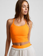 Woman wearing an orange cropped tank top and white bermuda shorts against a plain background.