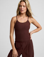 Woman wearing a brown waist length tank top and pants against a plain background.