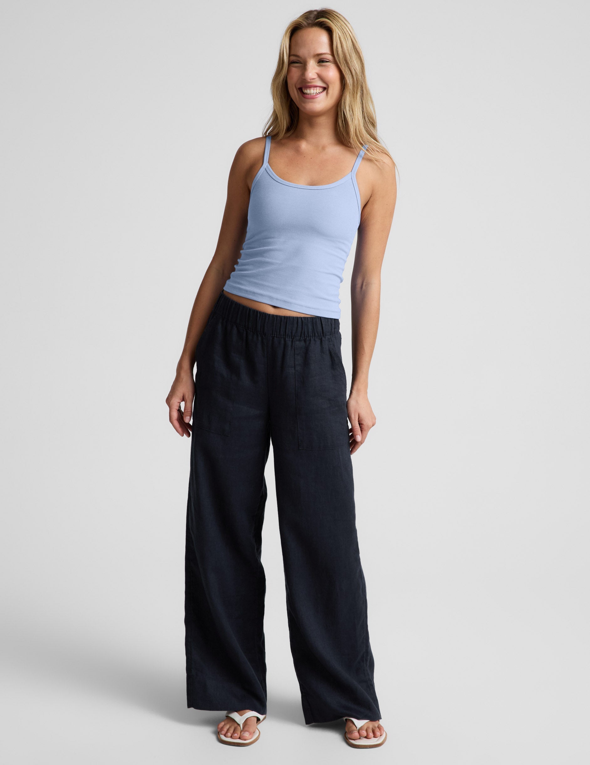 Woman wearing a light blue tank top and navy blue linen pants against a plain background.