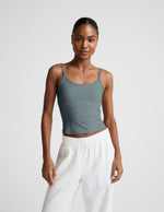 Woman wearing a green tank top and white pants against a plain background.