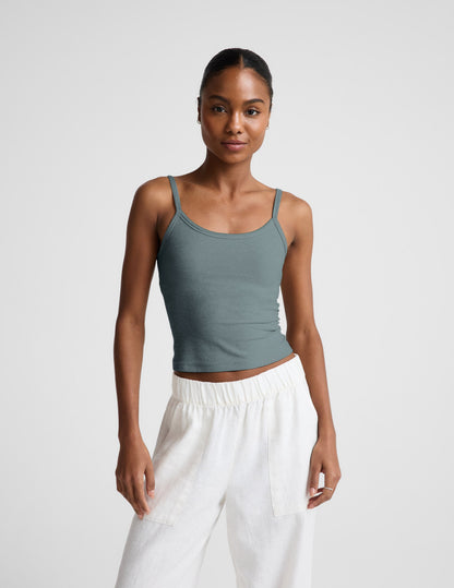 Woman wearing a green tank top and white pants against a plain background.