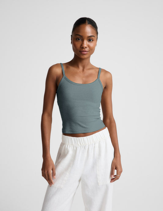 Woman wearing a green tank top and white pants against a plain background.
