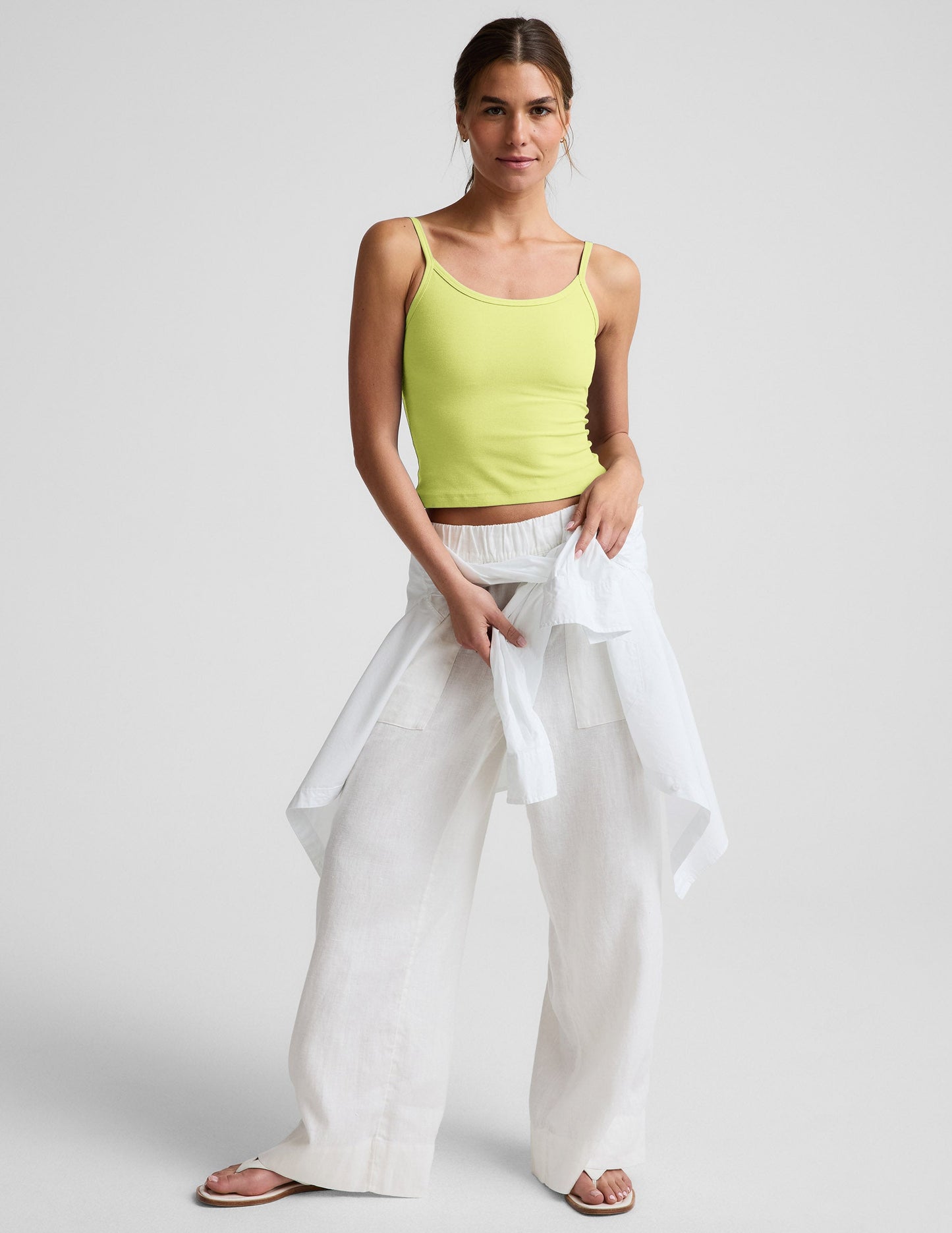 Woman wearing a yellow waist length tank and white linen pants against a plain background.