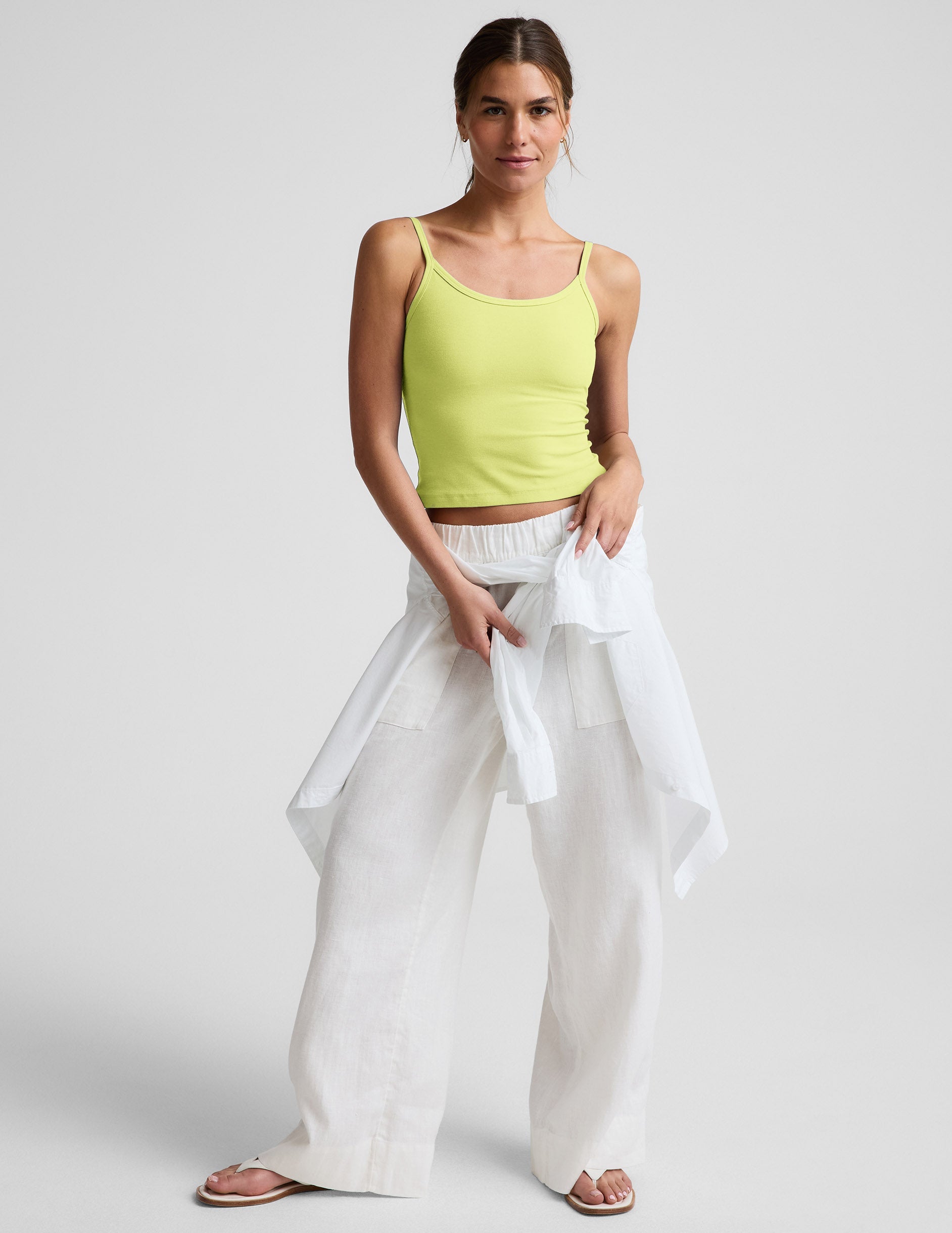 Woman wearing a yellow waist length tank and white linen pants against a plain background.