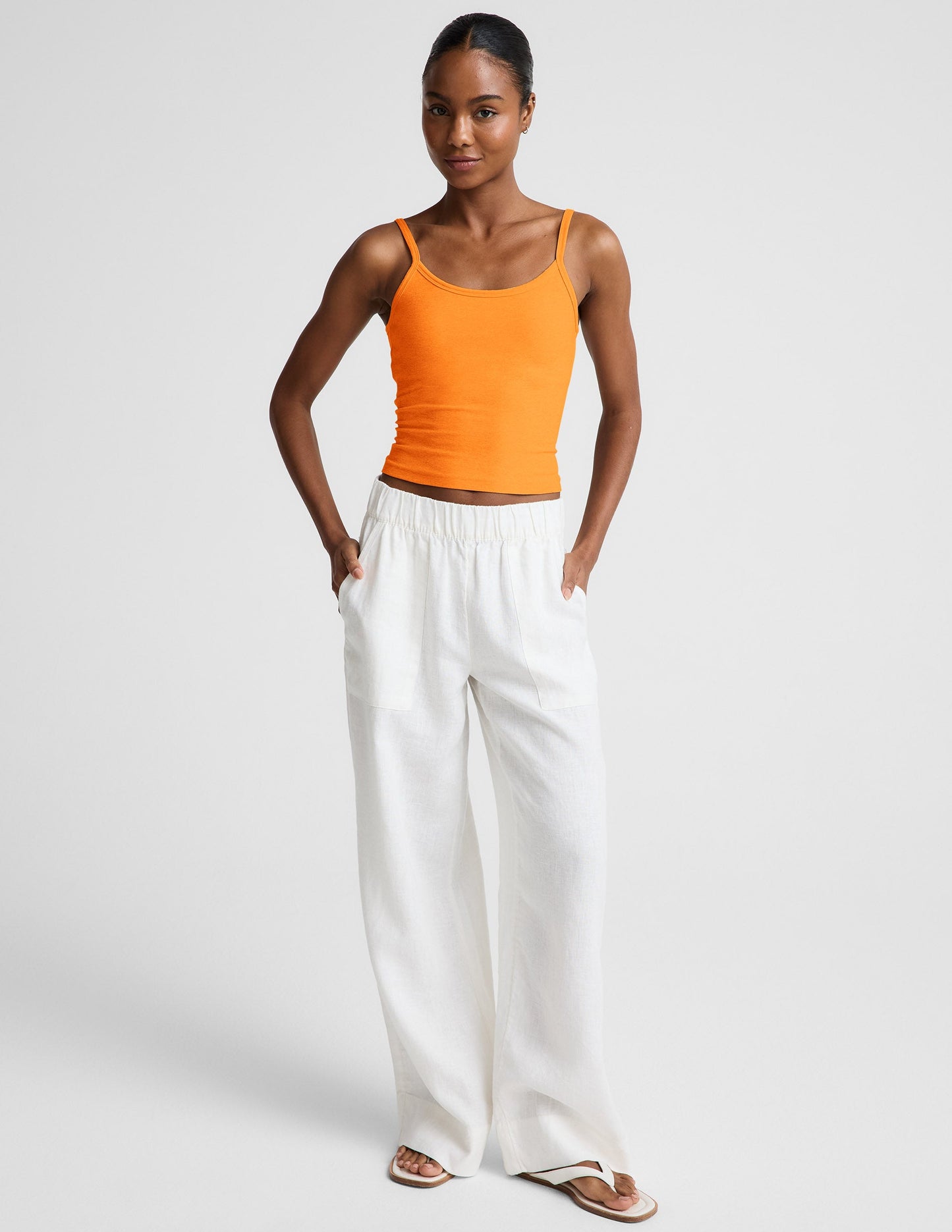 Woman wearing an orange tank top and white linen pants against a plain background.