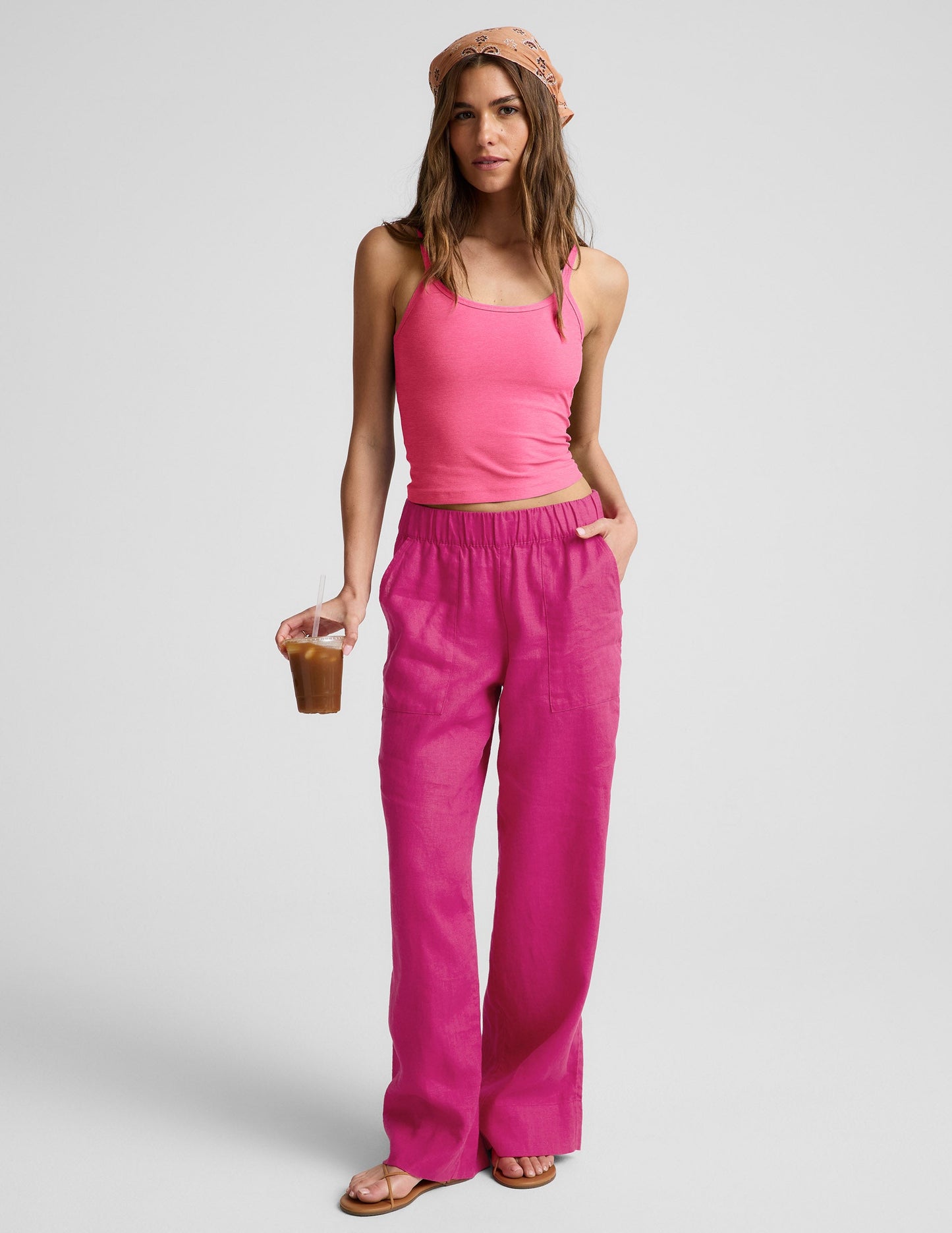 Woman wearing a pink waist length tank top and pink linen pants against a plain background.