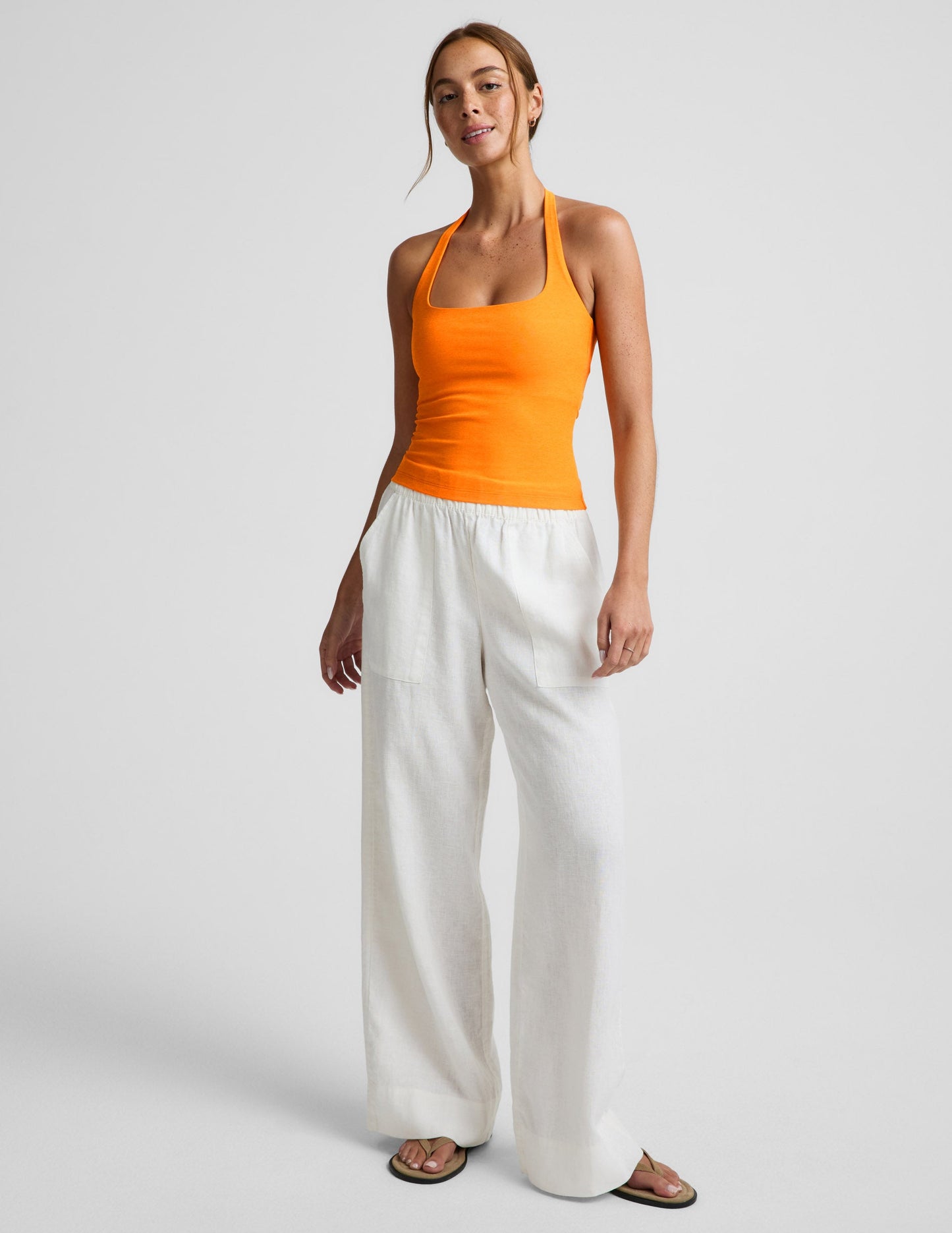 Woman wearing an orange halter tank top and white linen pants against a plain background.