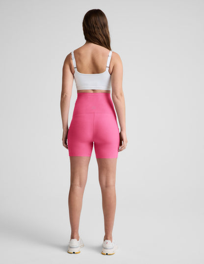 Woman wearing Pink Spacedye™ Keep Pace 5" Maternity Pocket Biker Short in a rear view with a white sports bra.