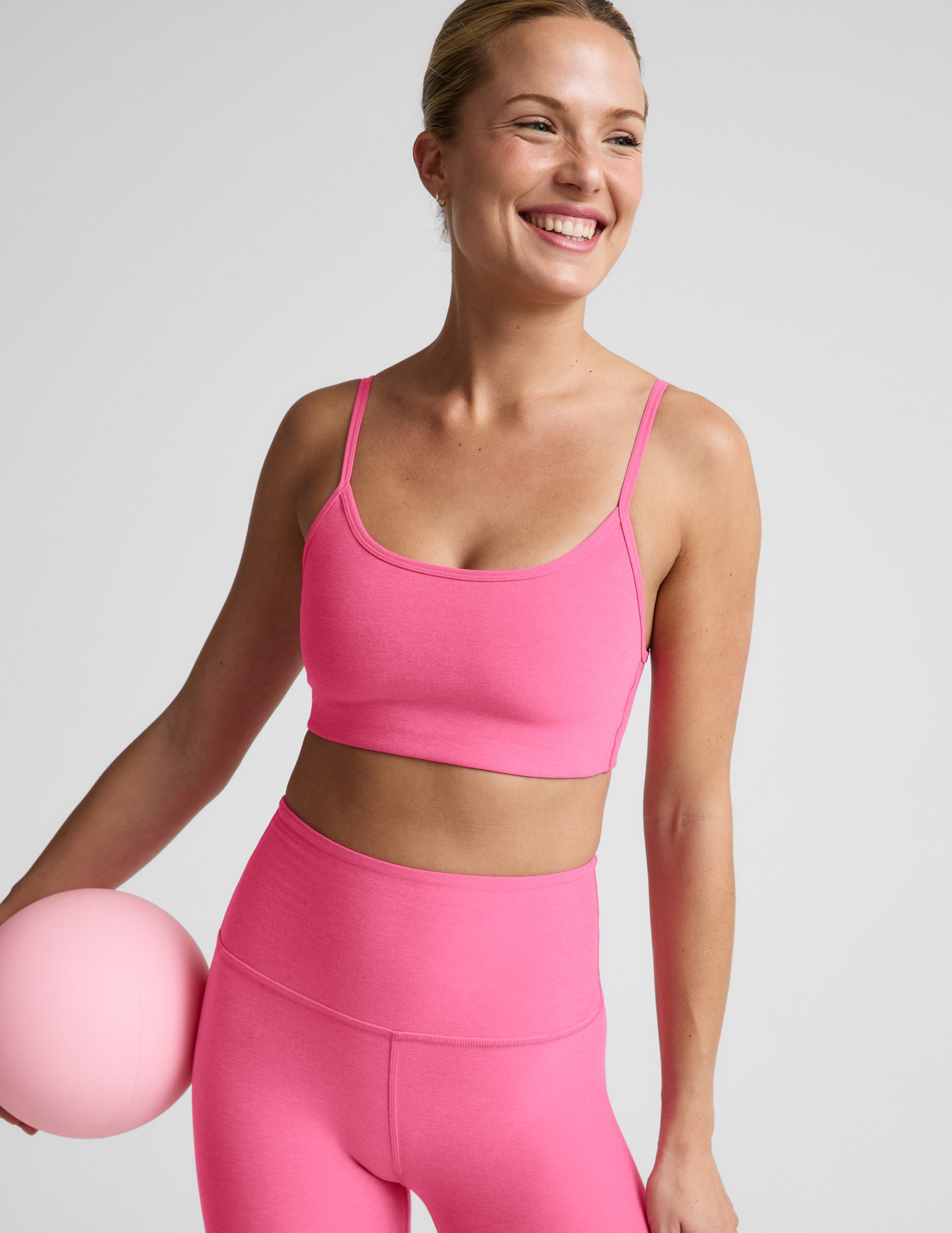 Woman wearing a pink sports bra and leggings holding a pink exercise ball against a light gray background.