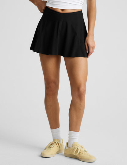 Woman wearing the Black You're A Force Skirt™ with a V-shaped waistline, paired with a white crop top and yellow sneakers.