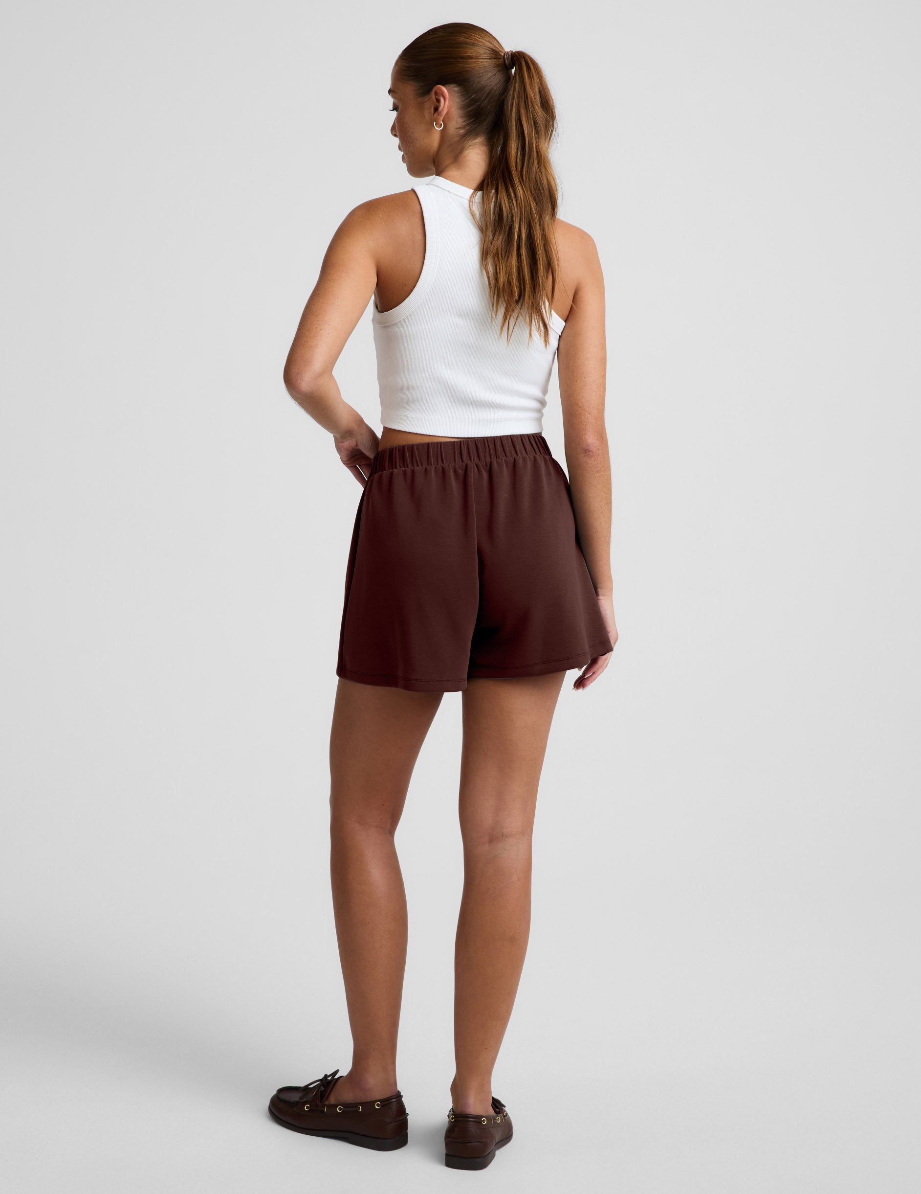 Woman wearing the brown Luxe Sandwash Skort™ with an elastic waistband, paired with a white tank top and loafers. Rear view.