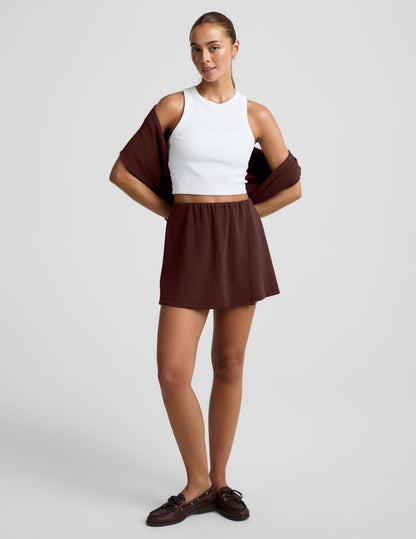 Woman wearing brown Luxe Sandwash™ Skort with a white ribbed tank top and a brown sweater draped over her shoulders.