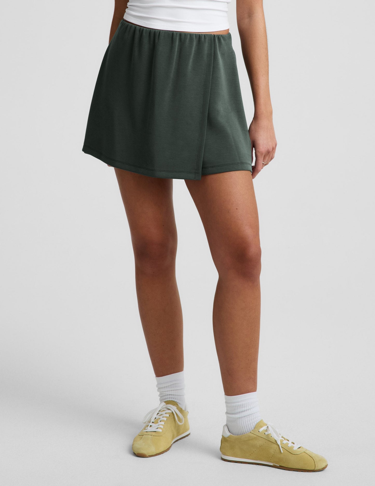 Woman wearing dark green Luxe Sandwash Skort with a wrap front, paired with a white tank top and yellow sneakers.