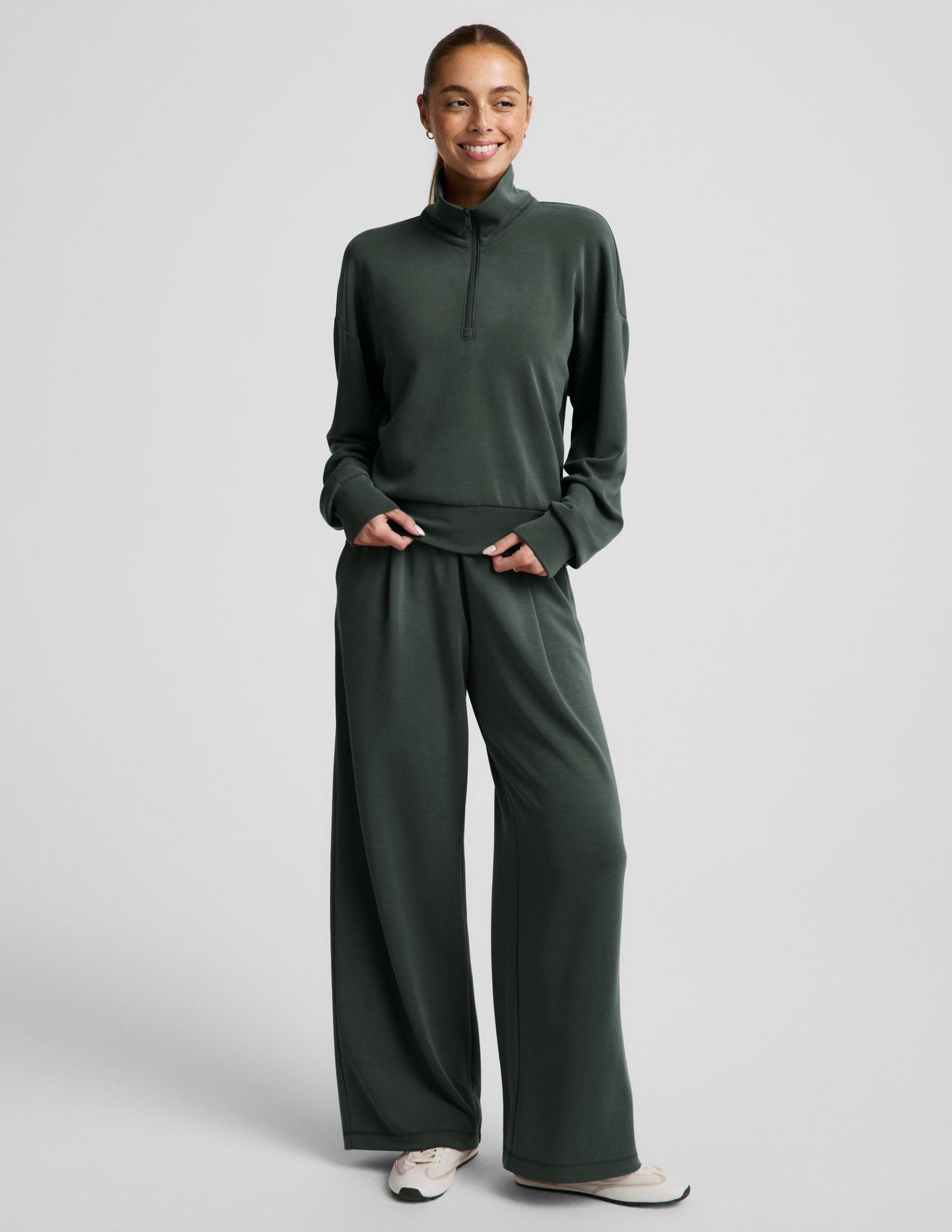 Woman wearing a green quarter zip sweatshirt and green wide leg pants against a plain background.