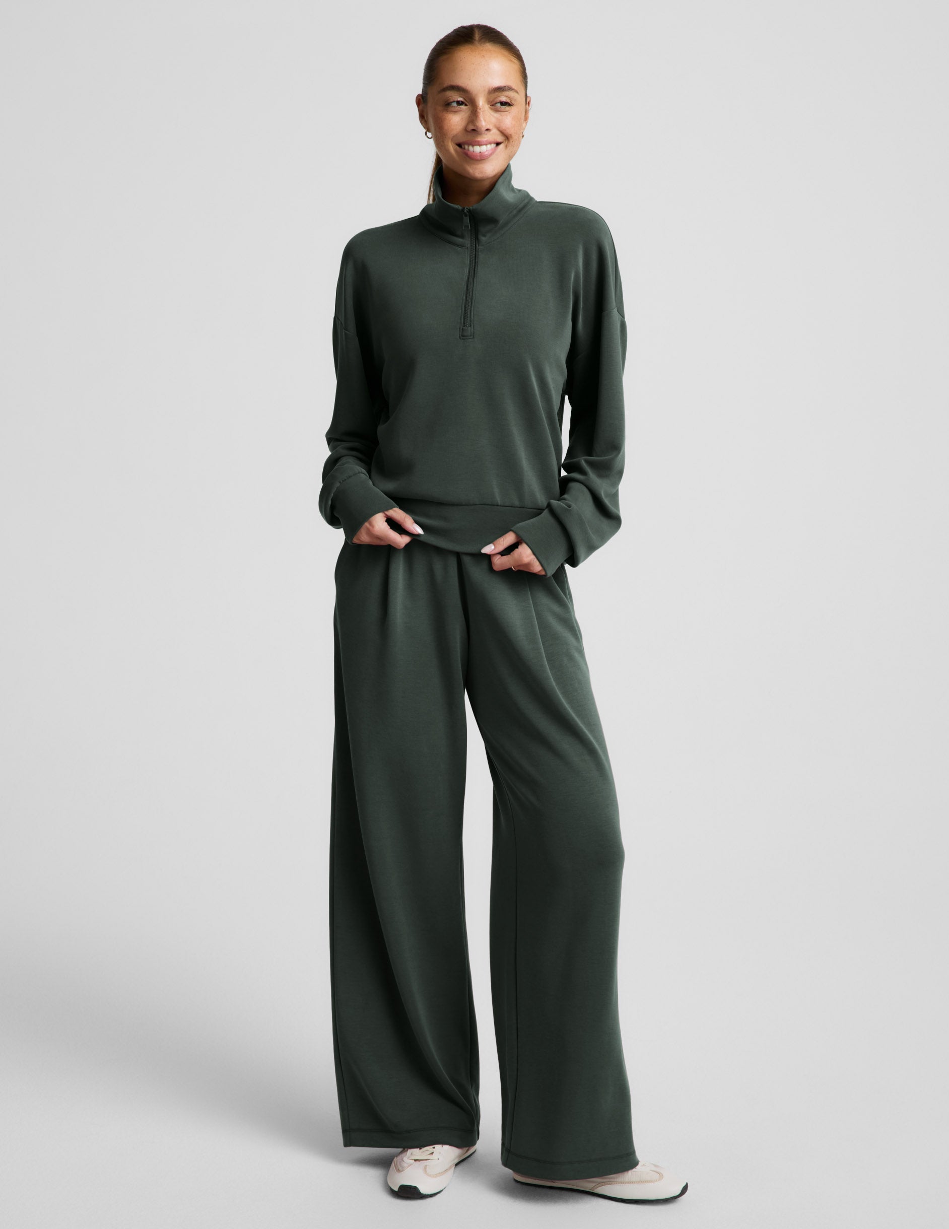 Woman wearing a green quarter zip sweatshirt and green wide leg pants against a plain background.