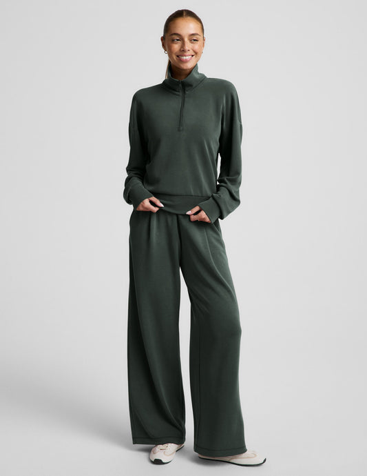 Woman wearing a green quarter zip sweatshirt and green wide leg pants against a plain background.