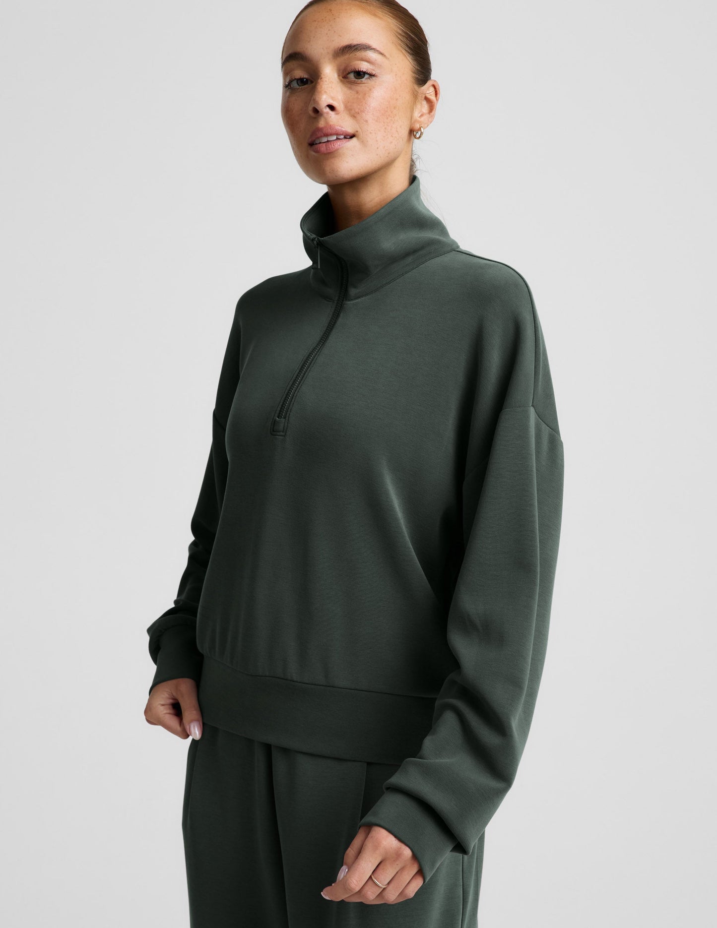 Woman wearing a green quarter zip sweatshirt and green wide leg pants against a plain background.