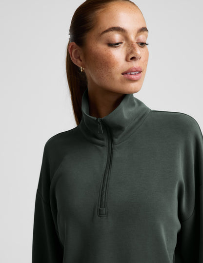 Woman wearing a green quarter zip sweatshirt and green wide leg pants against a plain background.
