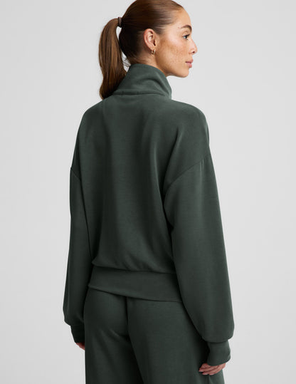 Woman wearing a green quarter zip sweatshirt and green wide leg pants against a plain background.