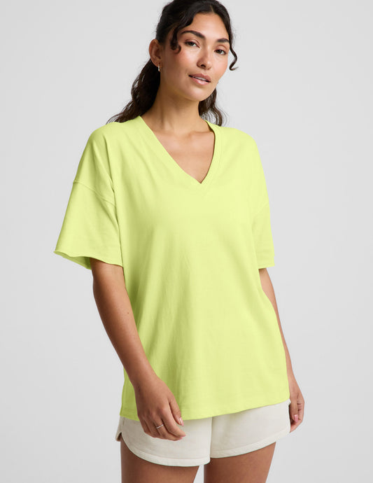 Woman wearing a bright level v-neck oversized t-shirt and white shorts against a plain background.