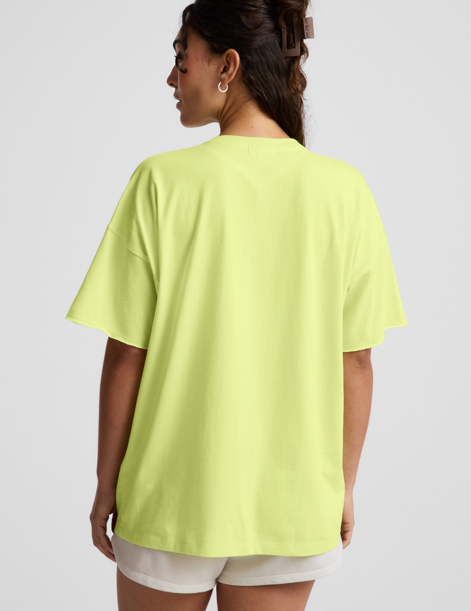 Woman wearing a bright level v-neck oversized t-shirt and white shorts against a plain background.