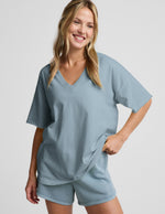 Woman wearing a blue v-neck oversized t-shirt and blue shorts against a plain background.