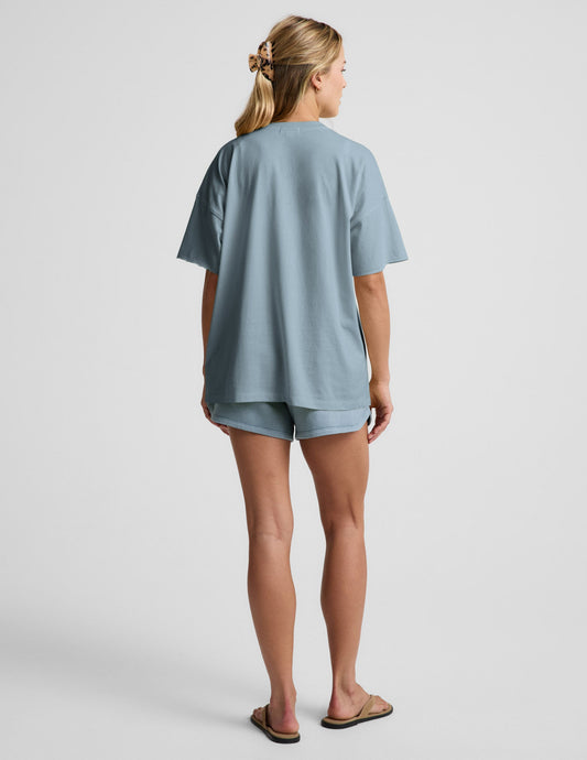 Woman wearing a blue v-neck oversized t-shirt and blue shorts against a plain background.