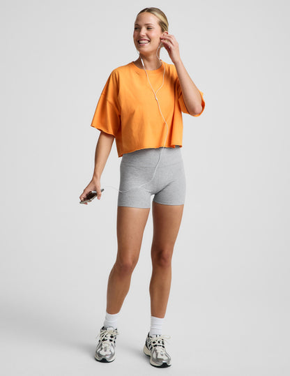 Woman wearing an orange cotton cropped t-shirt and gray biker shorts against a plain background.