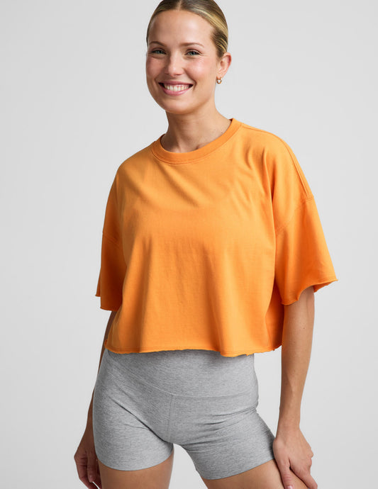 Woman wearing an orange cotton cropped t-shirt and gray biker shorts against a plain background.