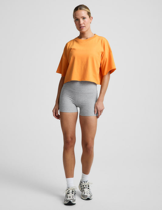 Woman wearing an orange cotton cropped t-shirt and gray biker shorts against a plain background.