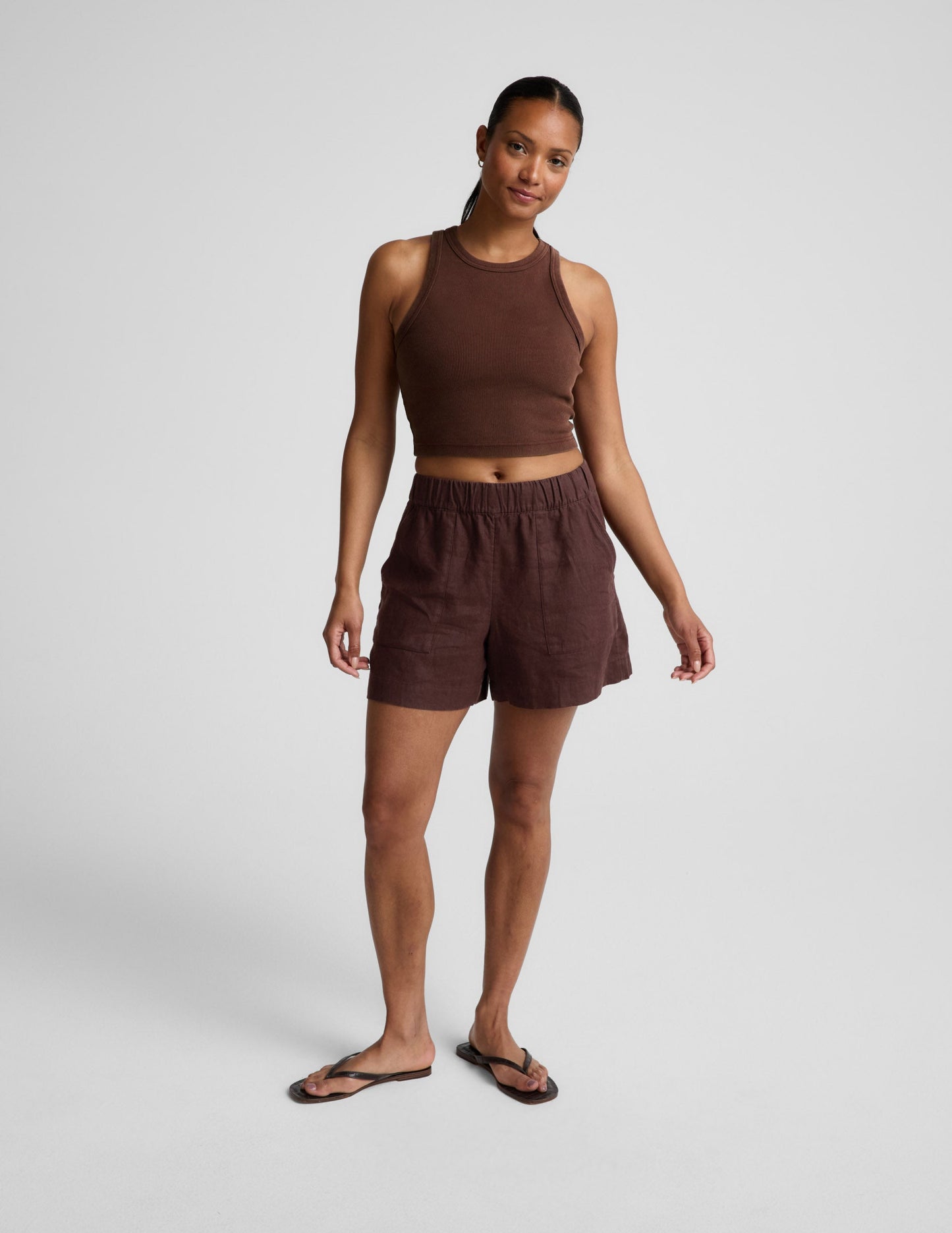 Woman wearing a brown ribbed cropped tank and brown linen shorts against a plain background.