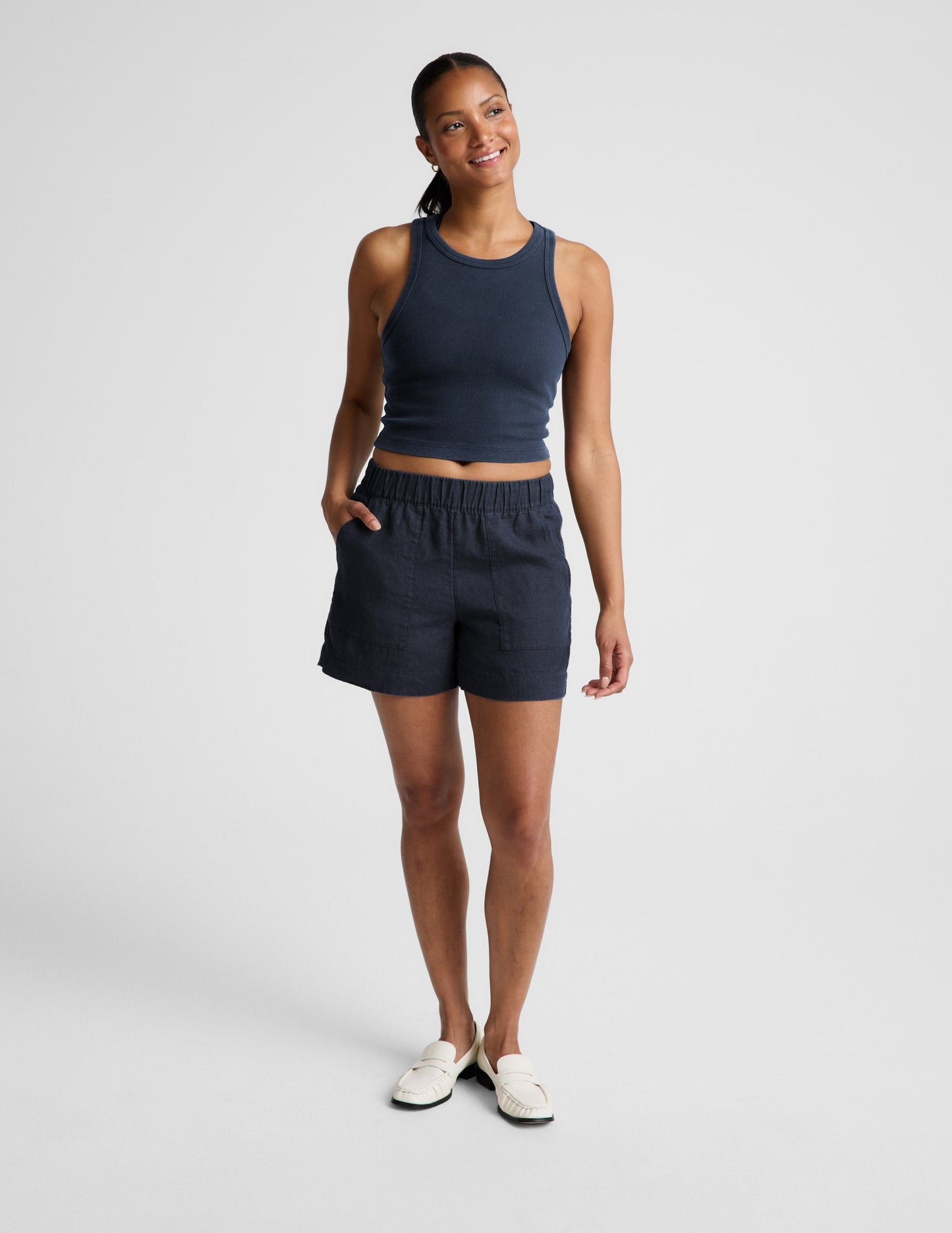 Woman wearing a navy ribbed tank top and linen shorts against a plain background.