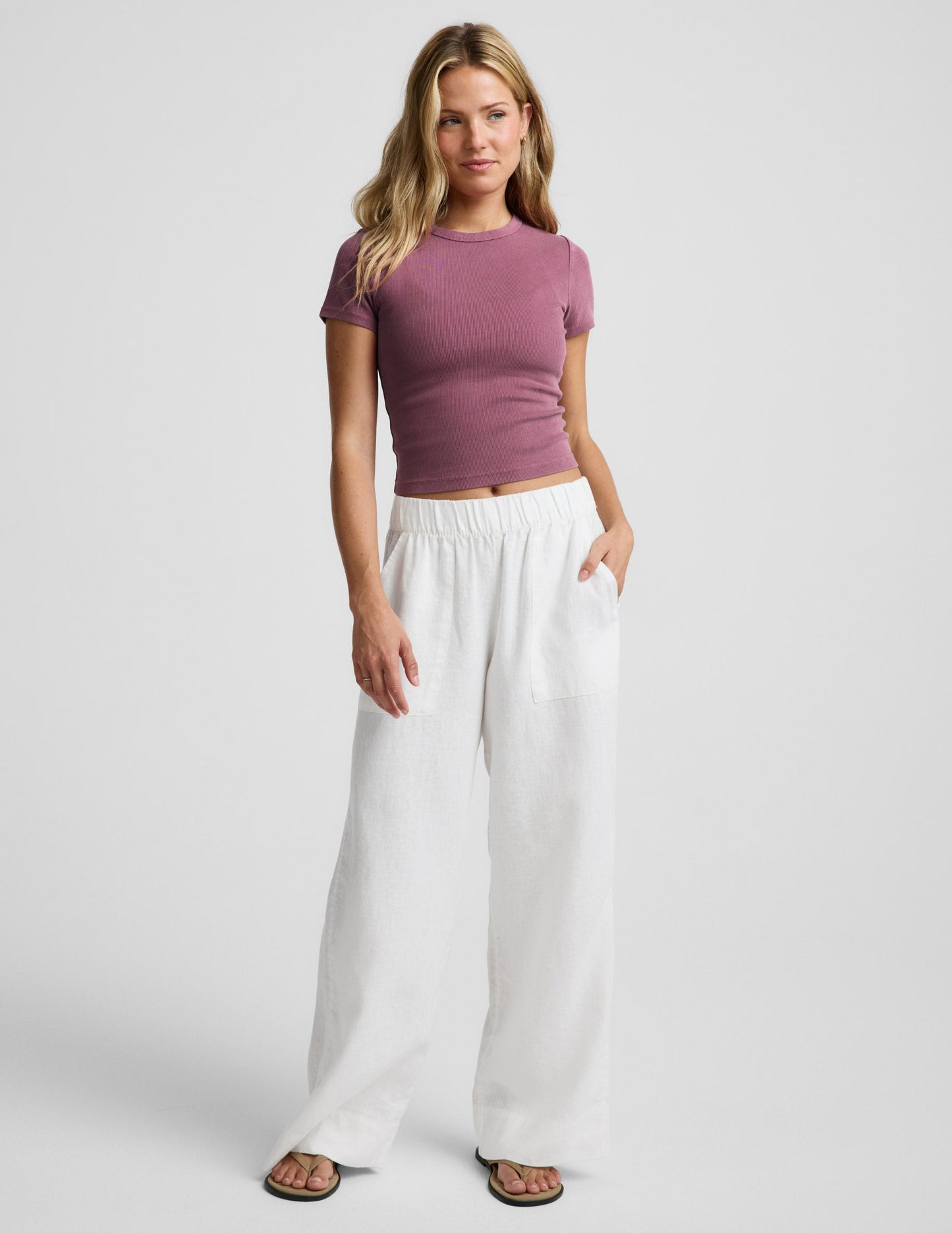 Woman wearing a purple ribbed baby tee and white linen pants against a plain background.