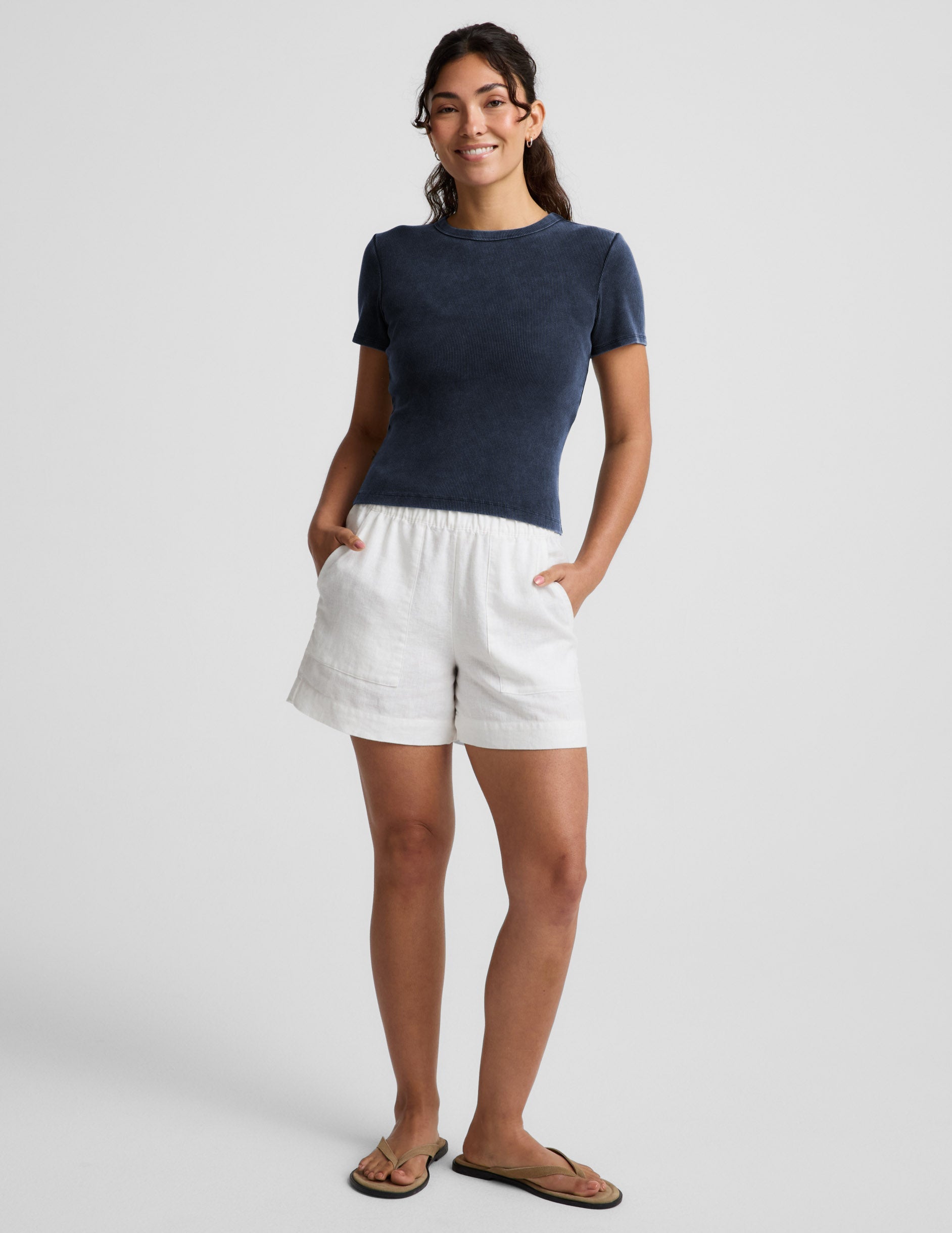 Woman wearing a navy blue ribbed t-shirt and white linen shorts against a plain background.
