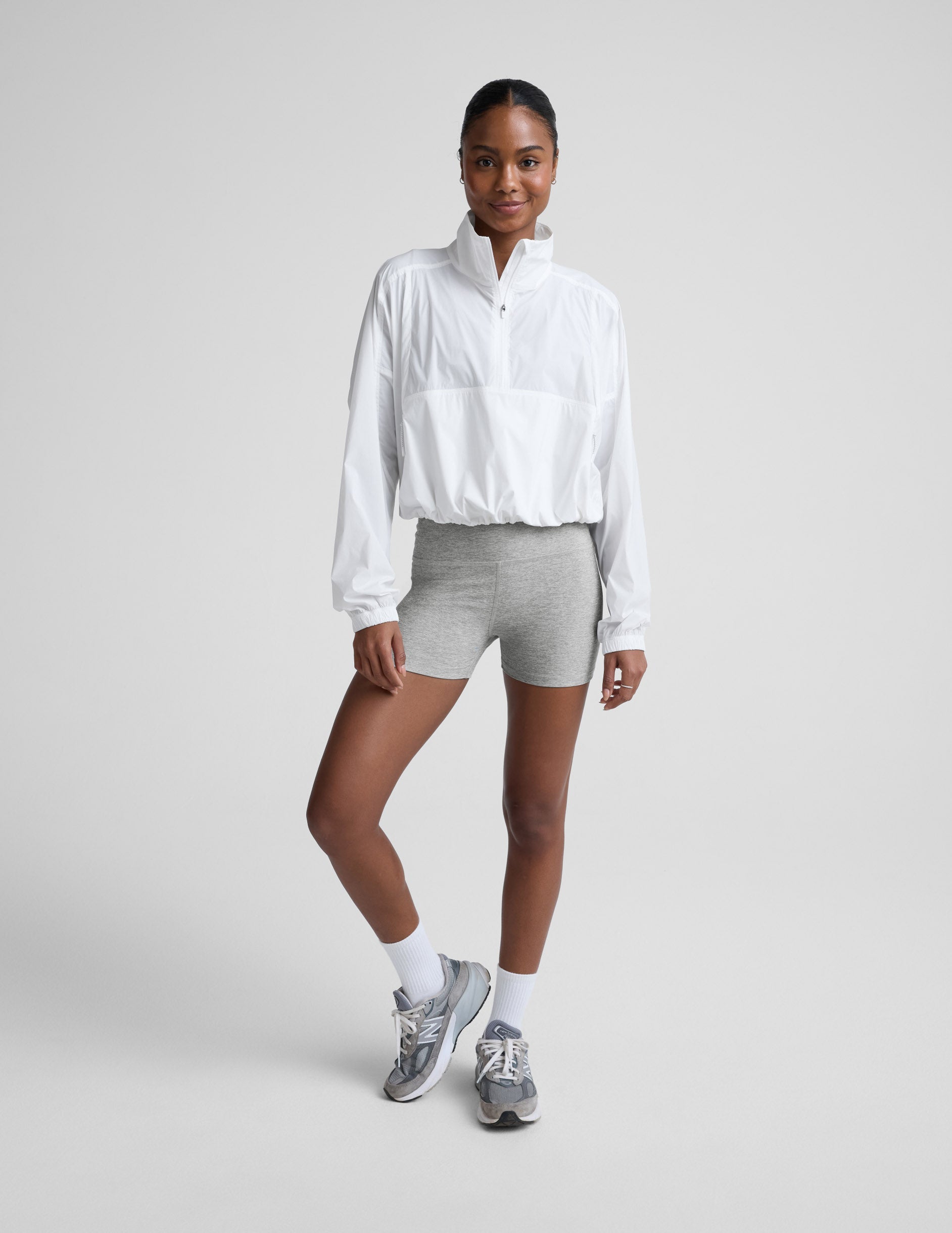 Woman wearing a white half-zip jacket and gray biker shorts against a plain background.