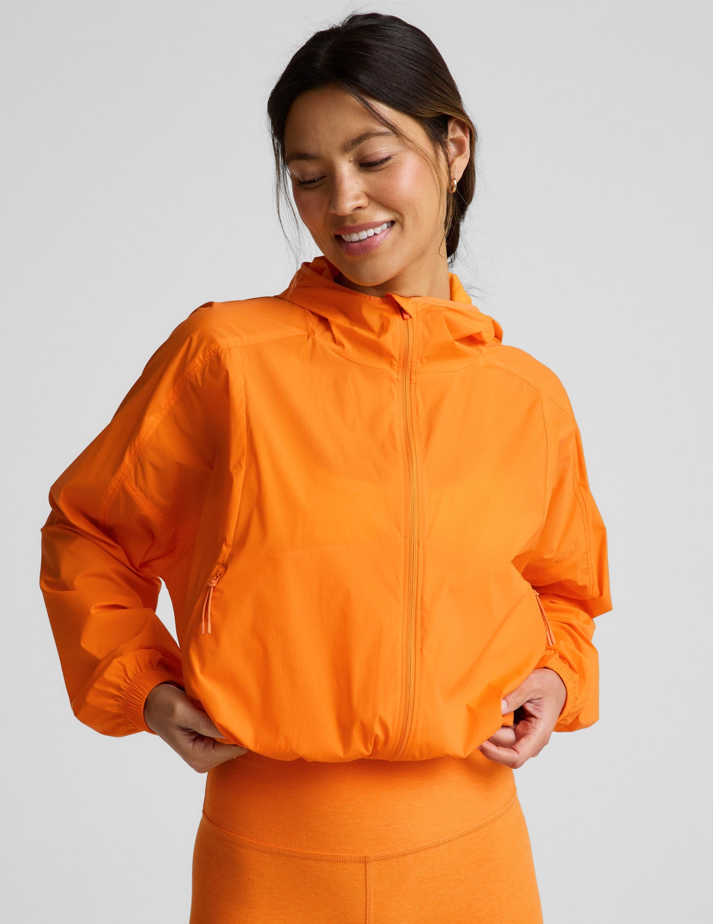 Woman wearing an orange jacket and orange biker shorts against a plain background.