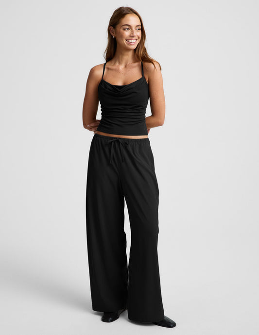 Woman wearing a black cowl neck tank top and black pants against a plain background.