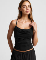 Woman wearing a black cowl neck tank top and black pants against a plain background.