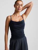 Woman wearing a navy cowl neck tank top and blue trousers against a plain background.