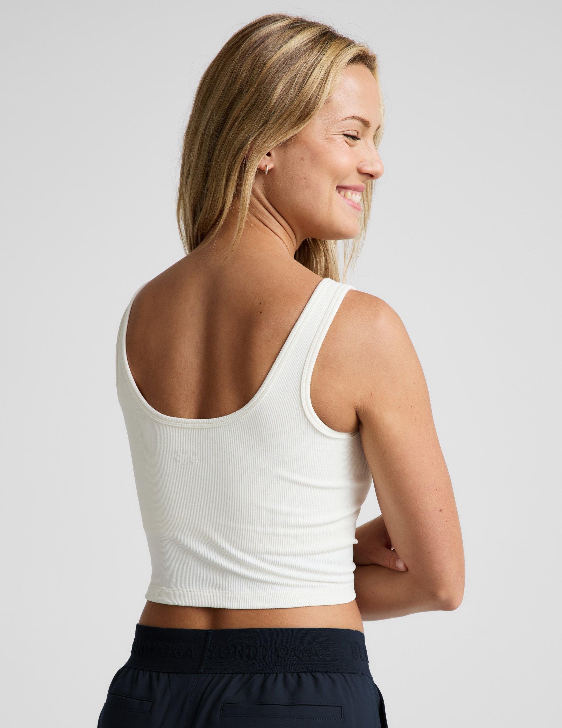 Woman wearing a white ribbed tank top and blue pants against a plain background.