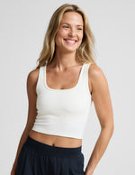 Woman wearing a white ribbed tank top and blue pants against a plain background.