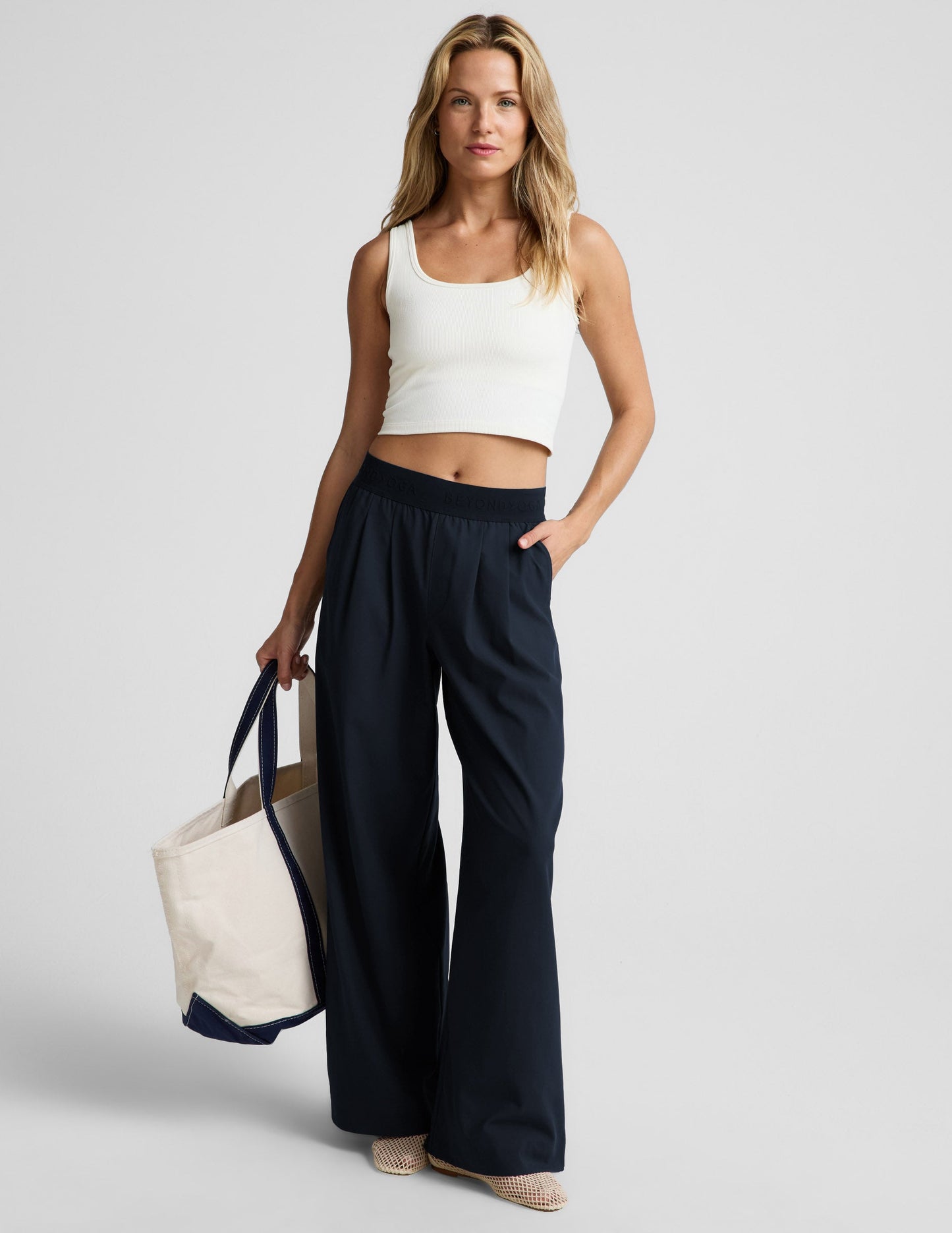 Woman wearing a white ribbed tank top and blue pants against a plain background.