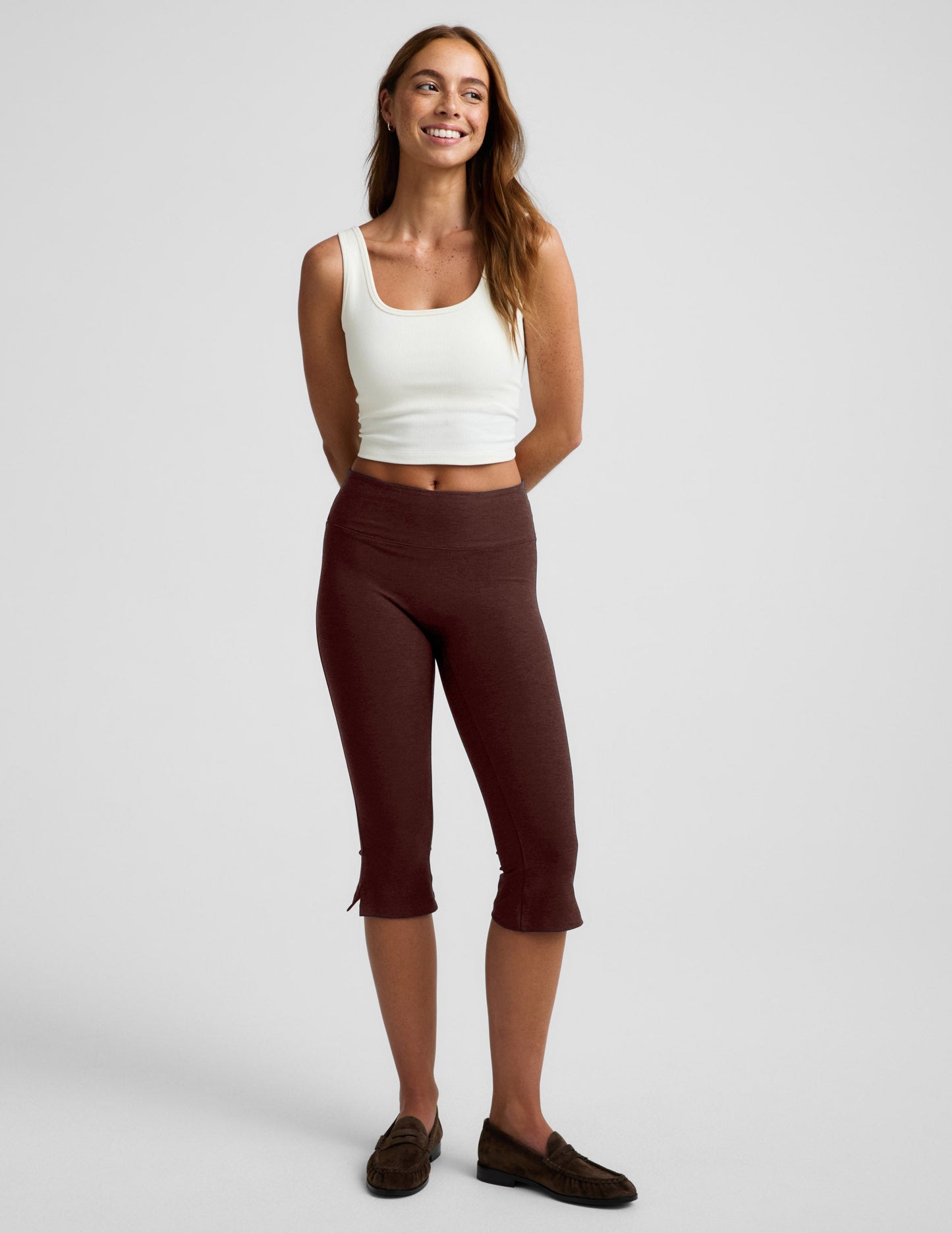 Woman wearing a white tank top and brown pedal pushers on a plain background.