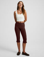 Woman wearing a white tank top and brown pedal pushers on a plain background.