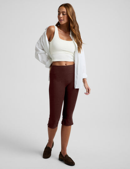 Woman wearing a white tank top and brown pedal pushers on a plain background.